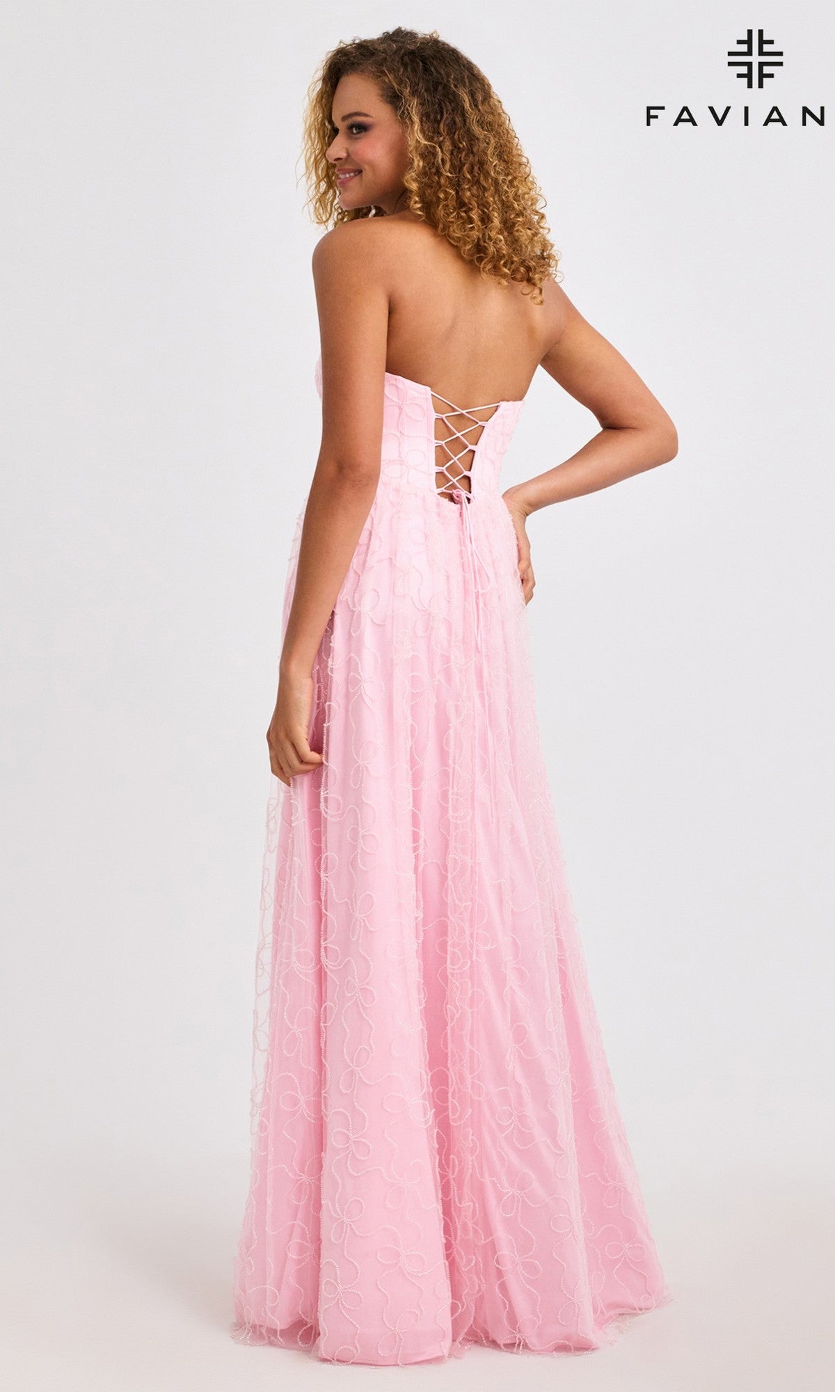 Wearing the Faviana 11418 long prom dress, a woman poses in a strapless pink gown with a corset bodice, lace-up back, beaded bow details, and a flowing textured skirt, looking over her shoulder as she stands facing away.